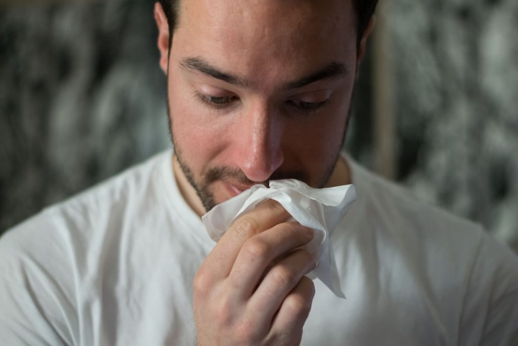 Person in a white shirt holding a tissue to their nose, appearing to sneeze, possibly due to allergies common in Dothan, AL.