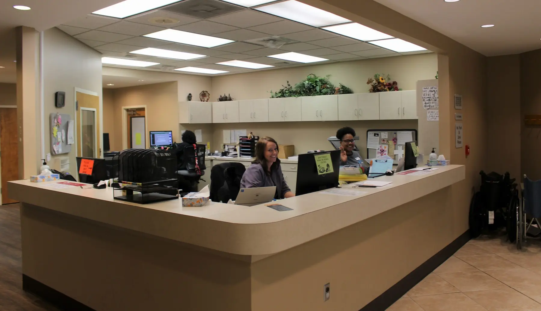 Two employees sit at a curved reception desk in an office setting with computers, paperwork, and office supplies visible.